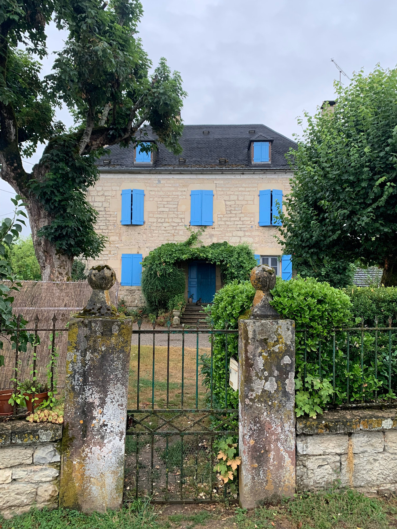 Une maison de pierre aux volets bleus derrière un portail et un jardin.